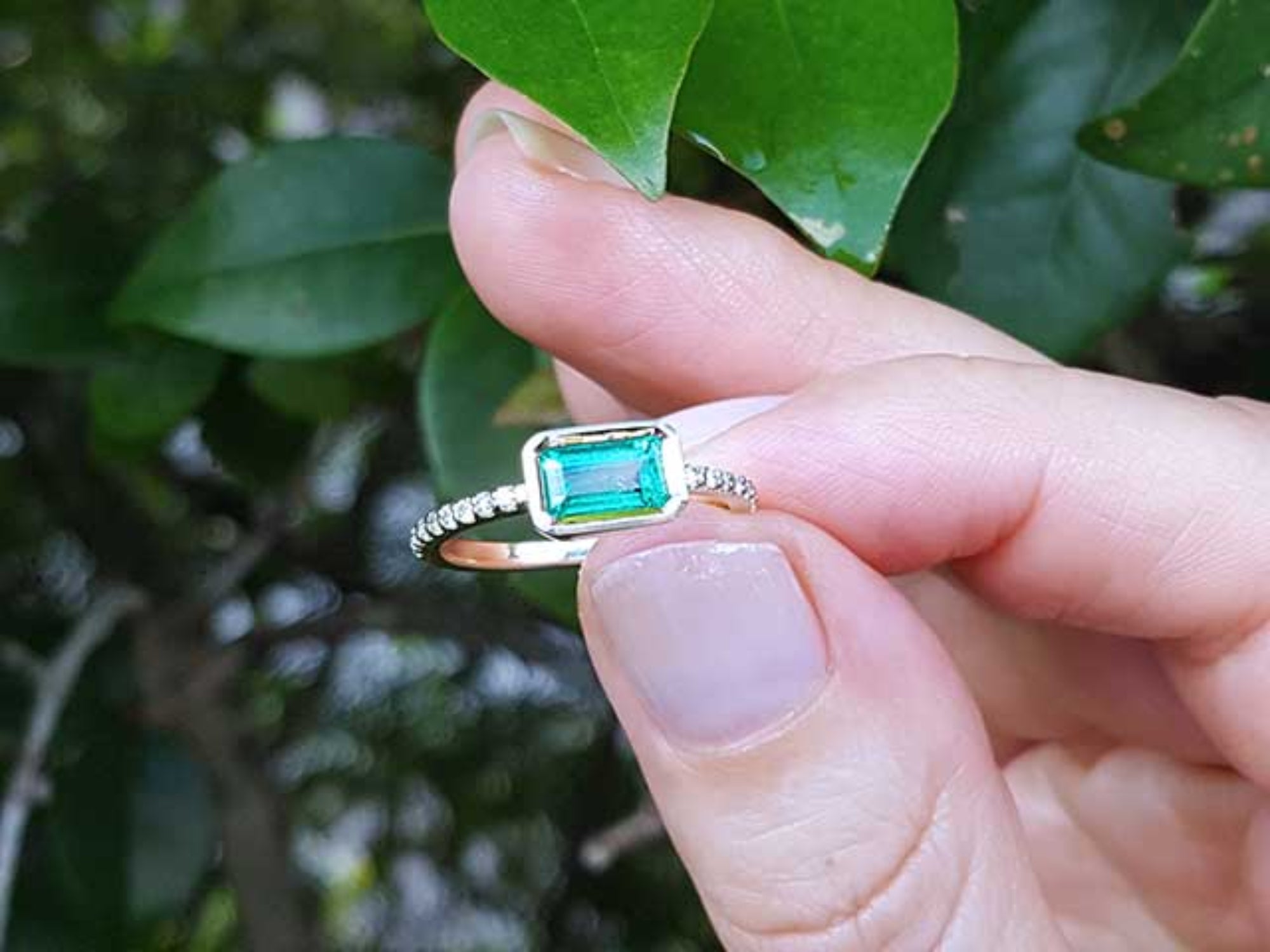A yellow gold emerald ring being held between fingers against a leafy background.