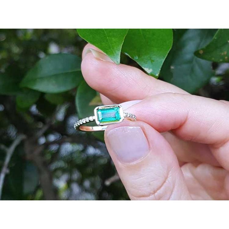 A yellow gold emerald ring being held between fingers against a leafy background.