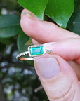 A yellow gold emerald ring being held between fingers against a leafy background.