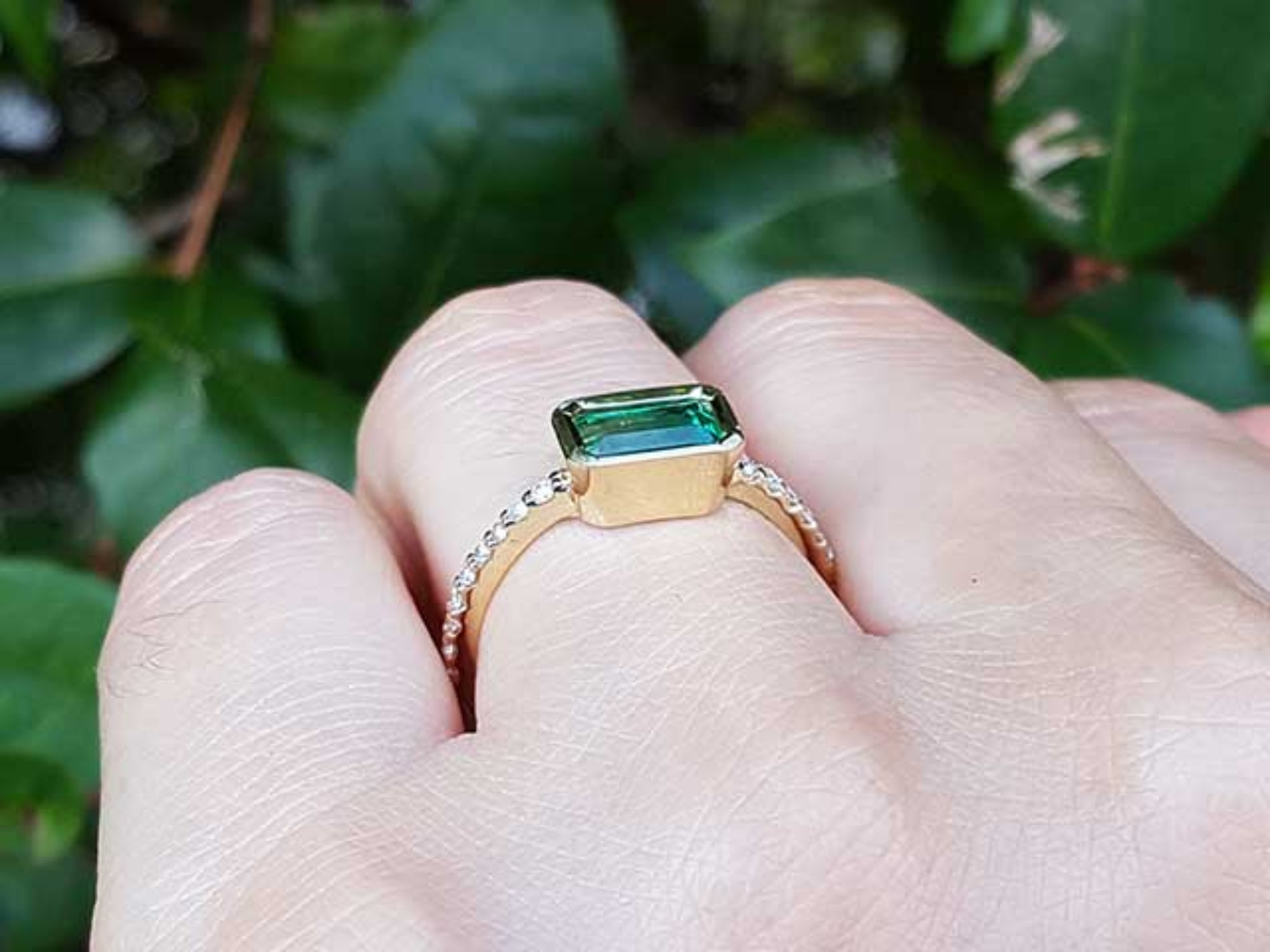 A yellow gold emerald ring on a hand against a green leafy background.