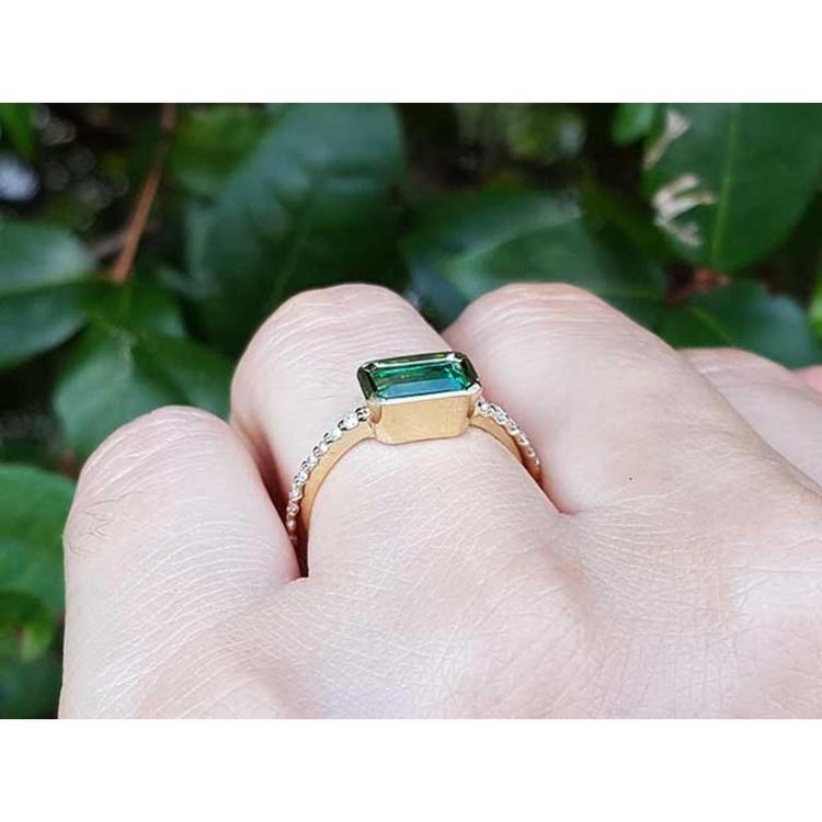 A yellow gold emerald ring on a hand against a green leafy background.