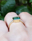 A yellow gold emerald ring on a hand against a green leafy background.