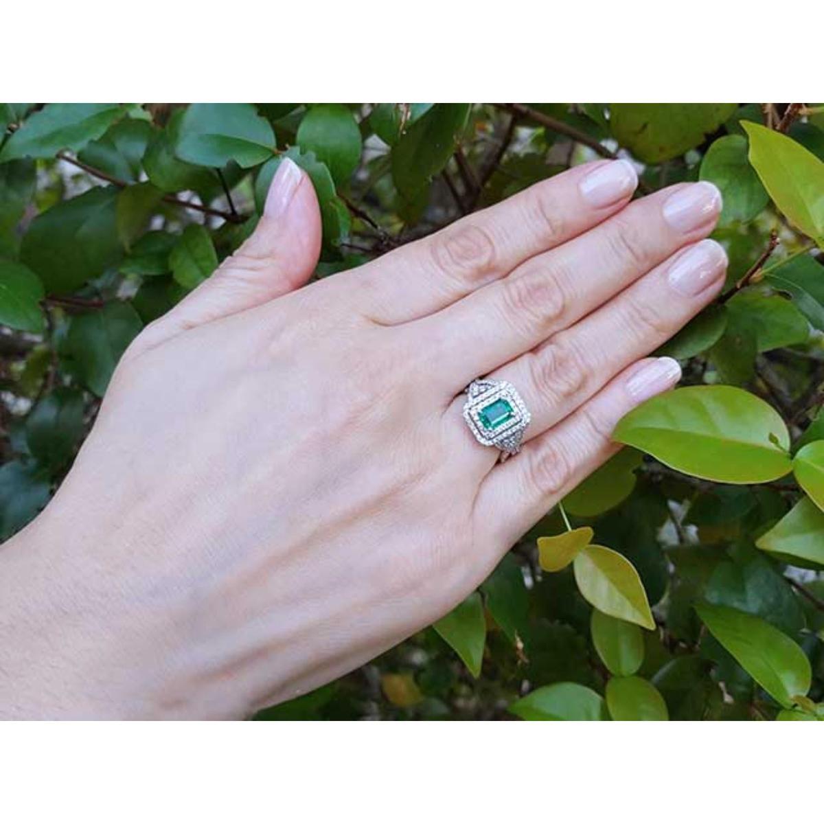 Hand wearing a ring with an emerald stone against a green leafy background.