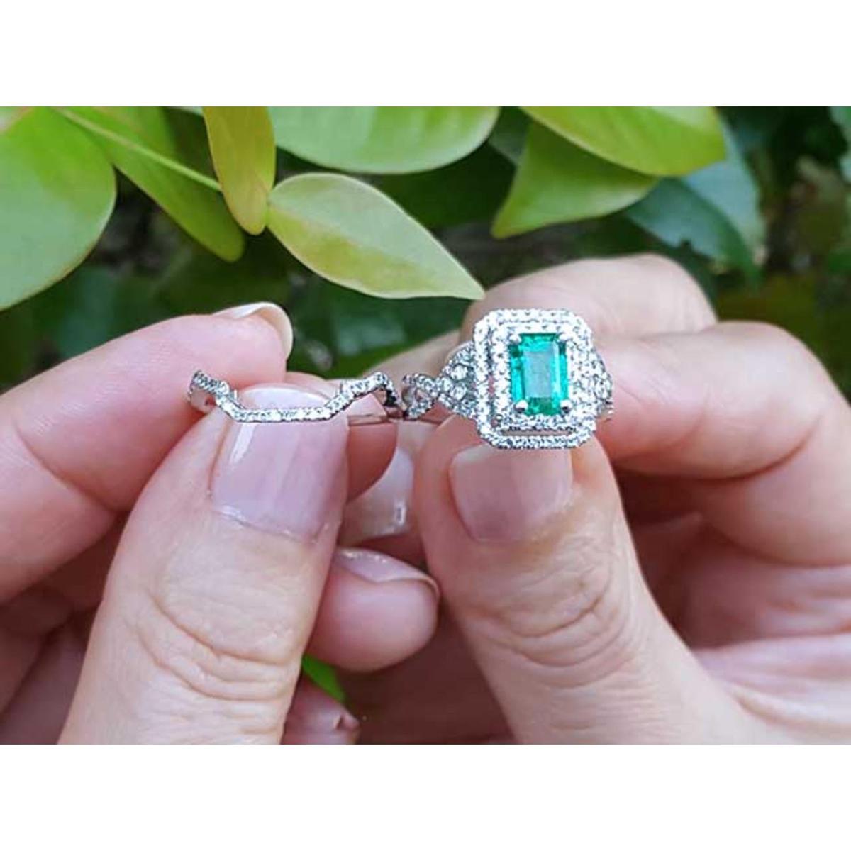 Close-up of a hand holding an emerald ring with matching band against a blurred green leafy background.