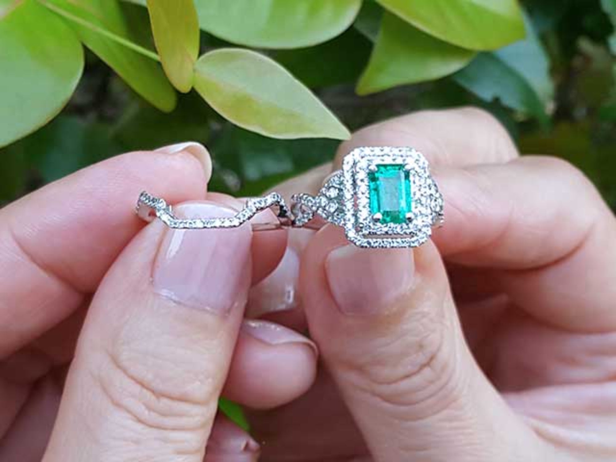 Close-up of a hand holding an emerald ring with matching band against a blurred green leafy background.