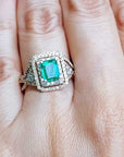 A bridal emerald engagement ring on a hand against a natural background.