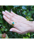 Hand wearing a yellow gold mens emerald ring against a green leafy background.
