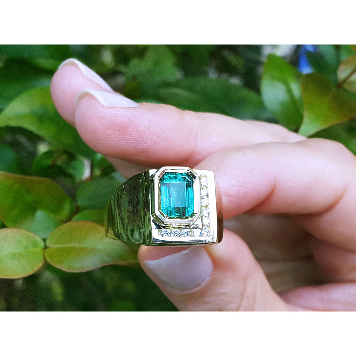 This rectangular emerald bezel set in a yellow gold ring for men is being held between fingers.