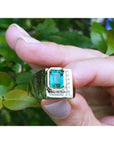 This rectangular emerald bezel set in a yellow gold ring for men is being held between fingers.