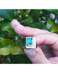 An emerald-cut men's emerald ring held by a hand between fingers against a green leafy background.