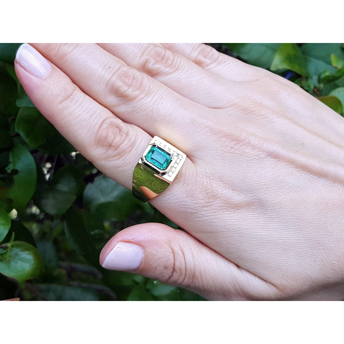 Gold ring for men with a colombian emerald on a hand against a green leafy background.