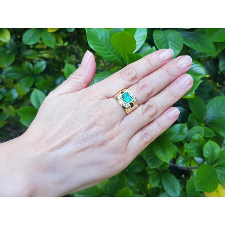 Hand wearing a gold men's emerald ring bezel set against a green leafy background.