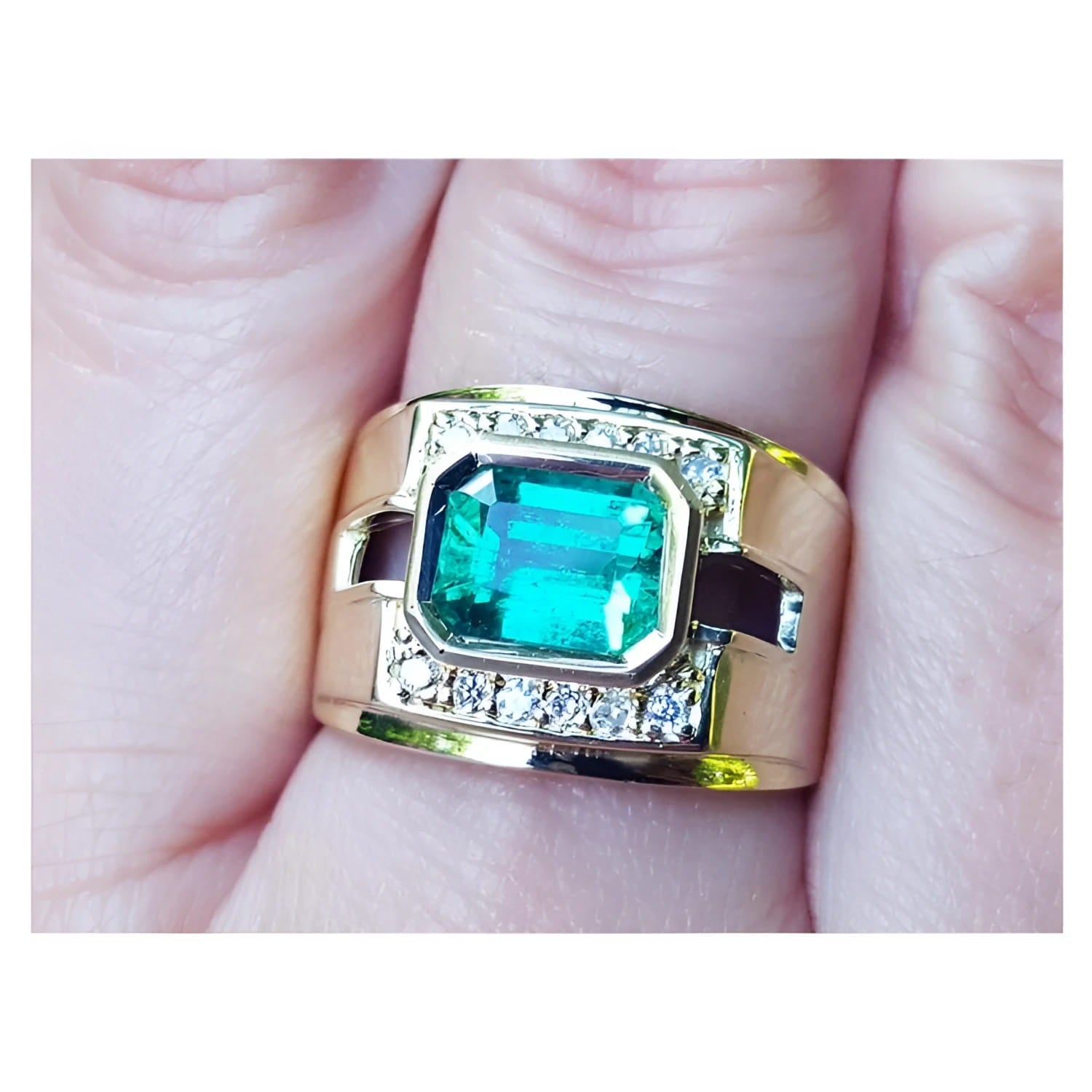 A 18k yellow gold natural emerald ring bezel set with side accent diamonds on a finger.