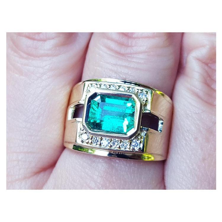 A 18k yellow gold natural emerald ring bezel set with side accent diamonds on a finger.