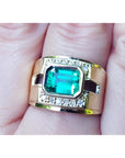 A 18k yellow gold natural emerald ring bezel set with side accent diamonds on a finger.