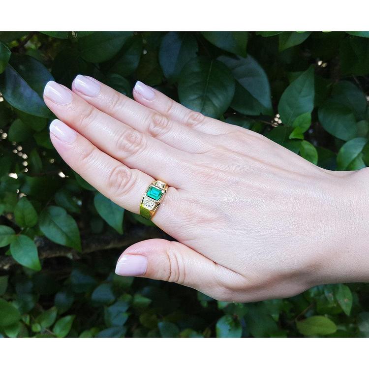 Hand wearing a yellow gold genuine emerald men's ring against a leafy background.