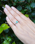 Hand wearing a ring with an emerald stone against a green leafy background.