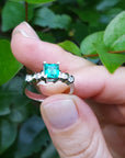 Hand holding a natural emerald ring against a green leafy background.