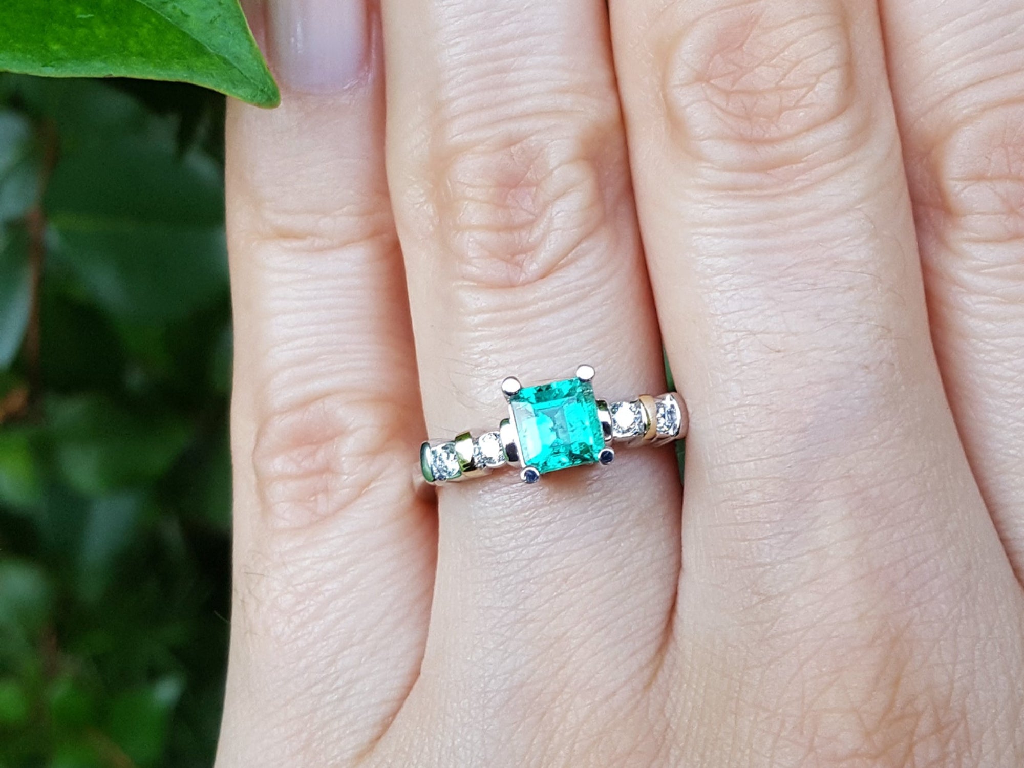 Close-up of a hand wearing a ring with a square emerald against a blurred green background.