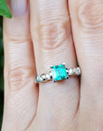 Close-up of a hand wearing a ring with a square emerald against a blurred green background.