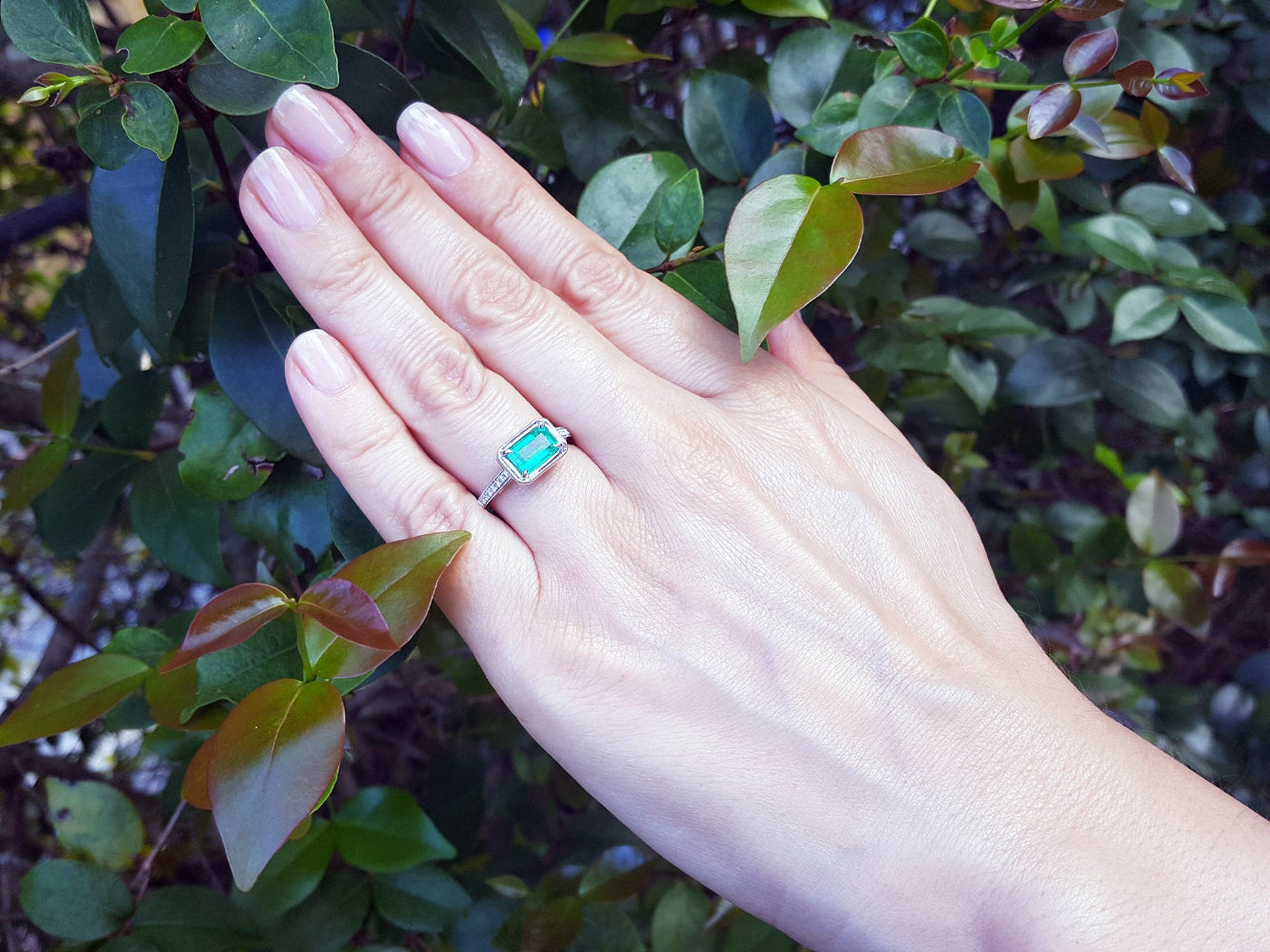 Hand wearing a natural emerald ring with accent diamonds against a green leafy background.