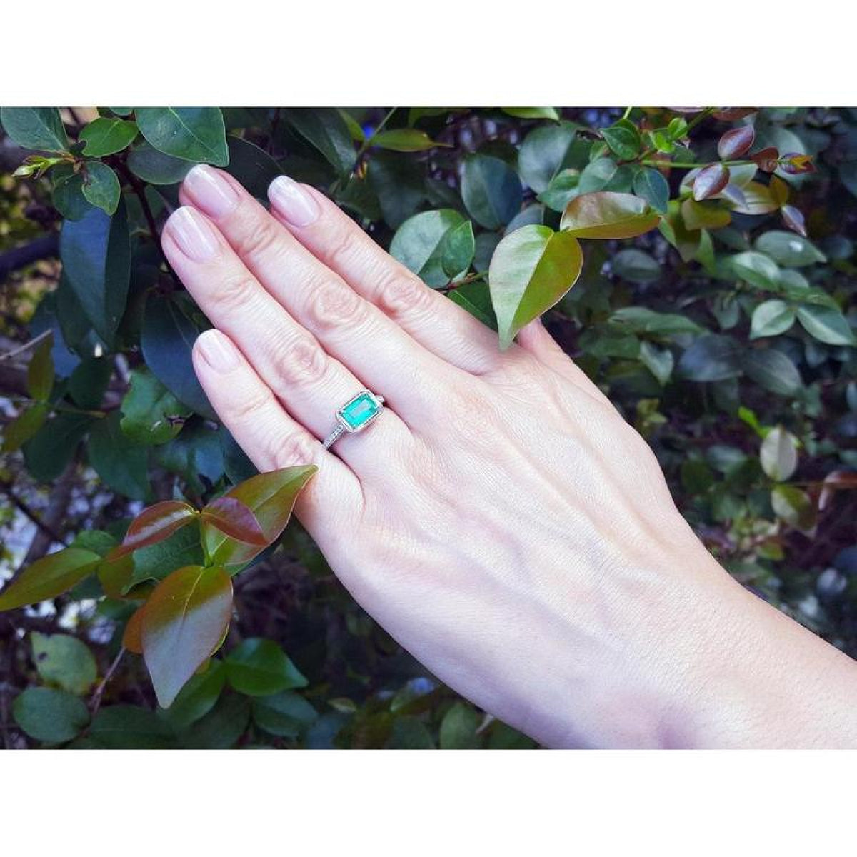 Emerald-cut emerald ring with natural diamonds on a 14K white gold band, worn on a hand amidst green leaves.