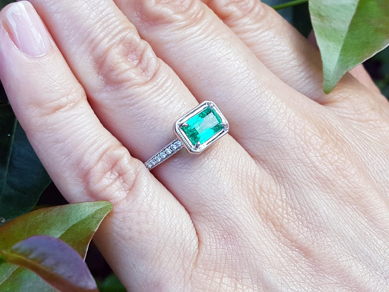 Emerald ring on a hand with green leaves in the background.