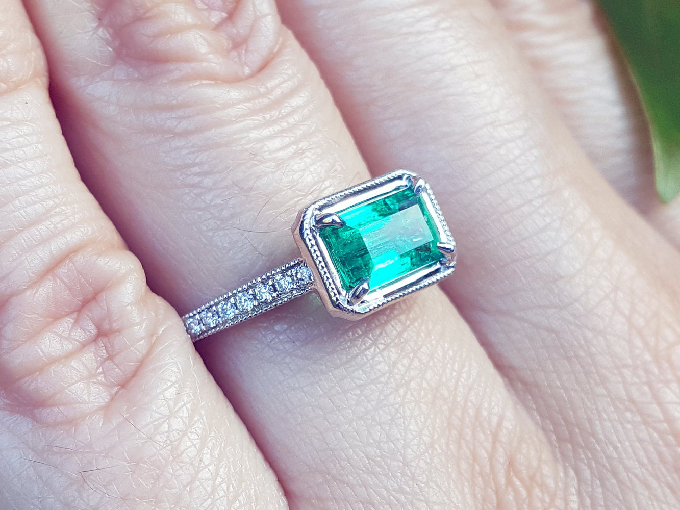 A white gold emerald ring with accent natural diamonds on a finger.