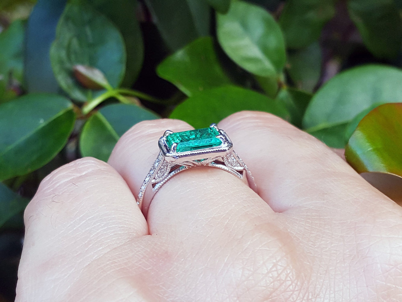 Ring with natural emerald on a finger against a leafy background.