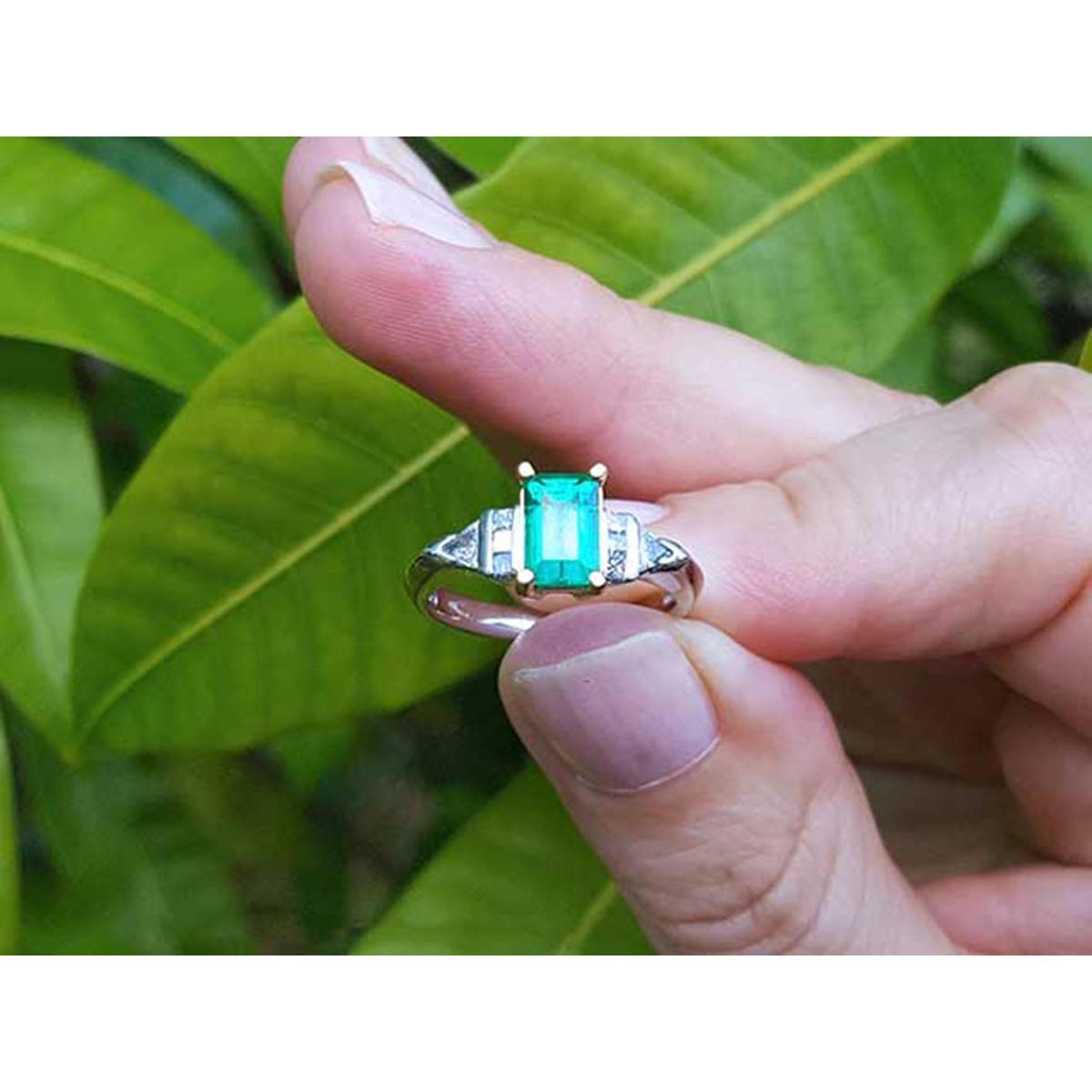 A platinum emerald ring being held between fingers against a leafy background.