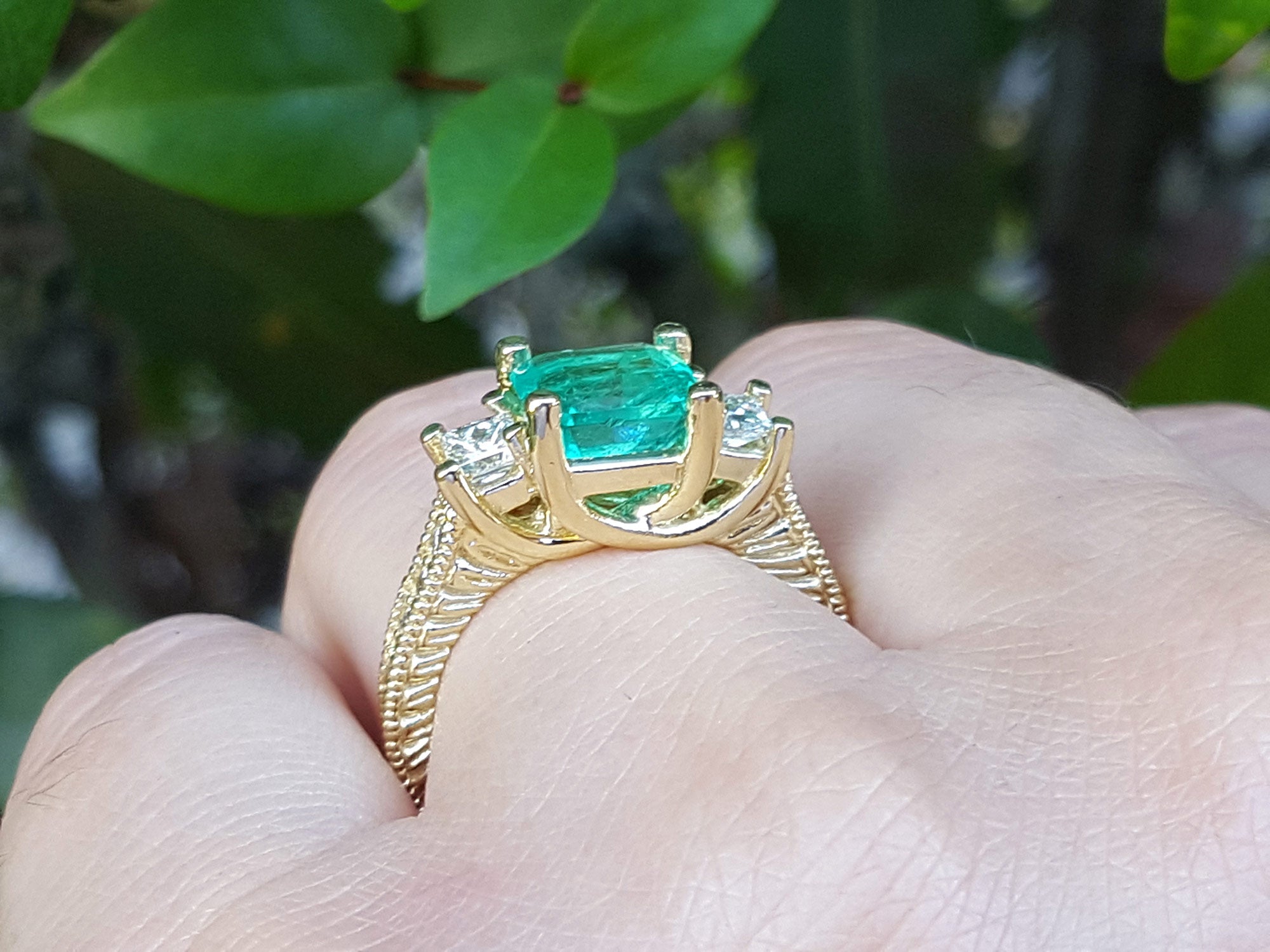 A Brial&#39;s may birthstone emerald ring held in a hand against a blurred green leafy background.