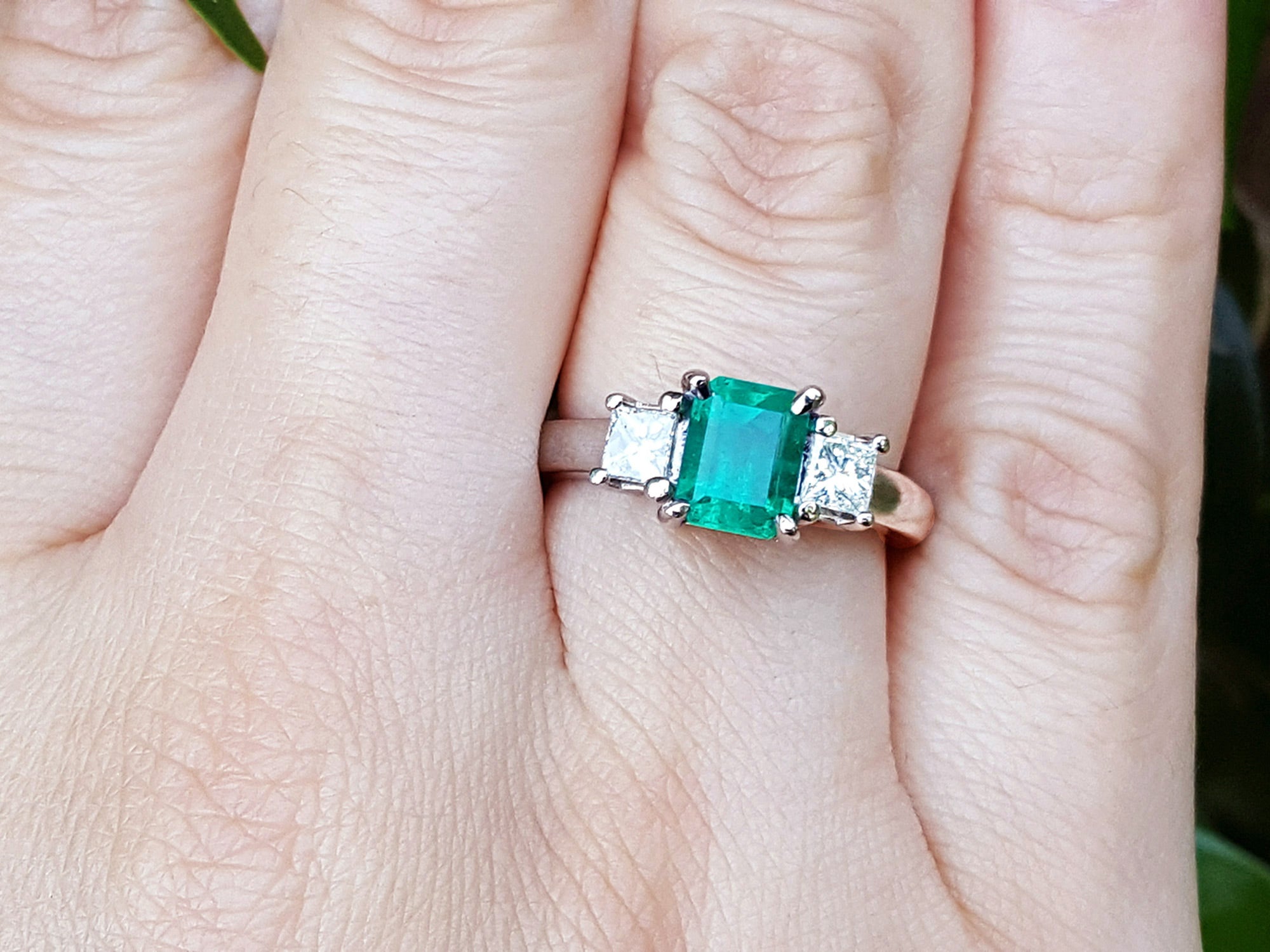 This three stone natural emerald ring with side princess cut diamonds is on a finger.