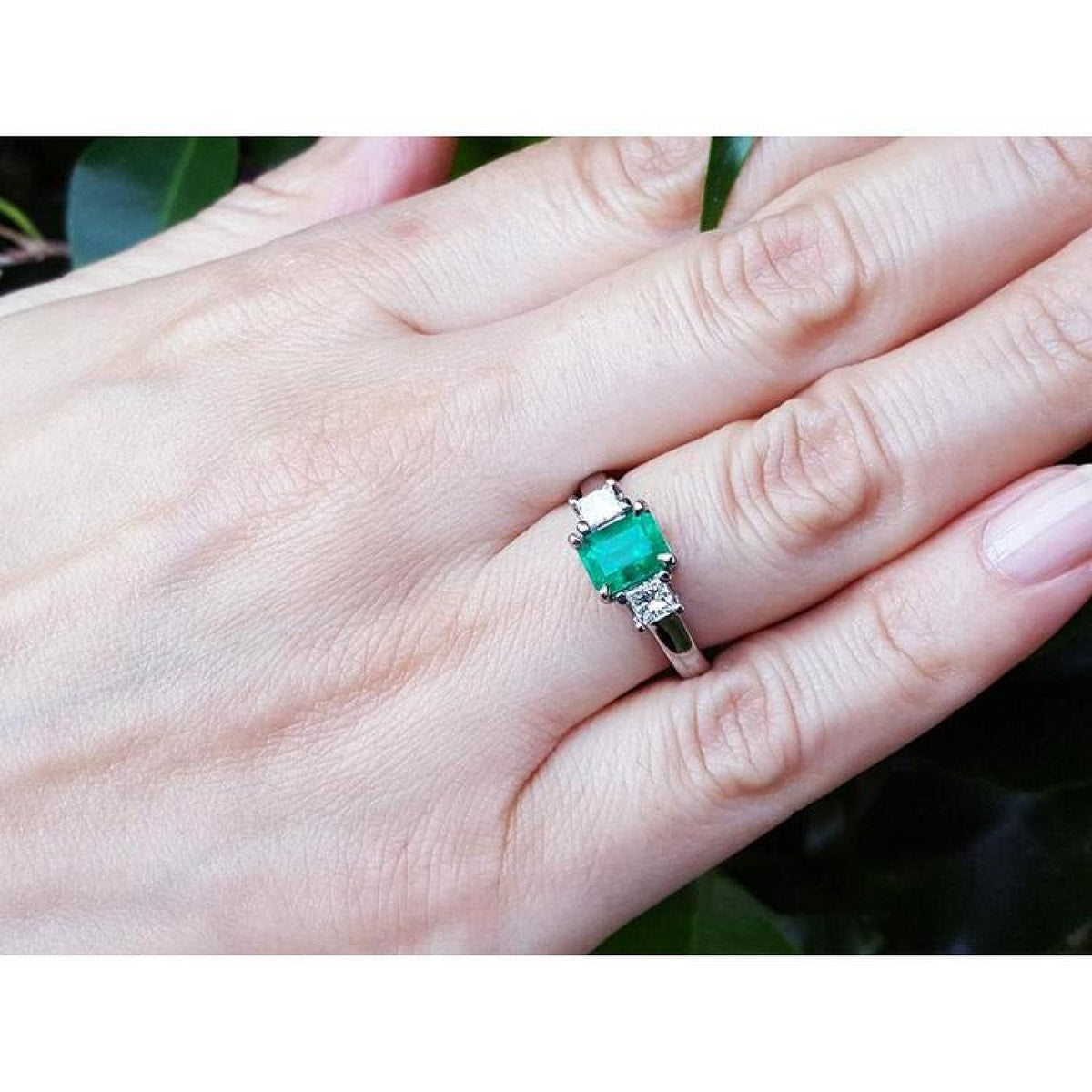 18k white gold three-stone emerald ring with princess cut side diamonds on a hand.