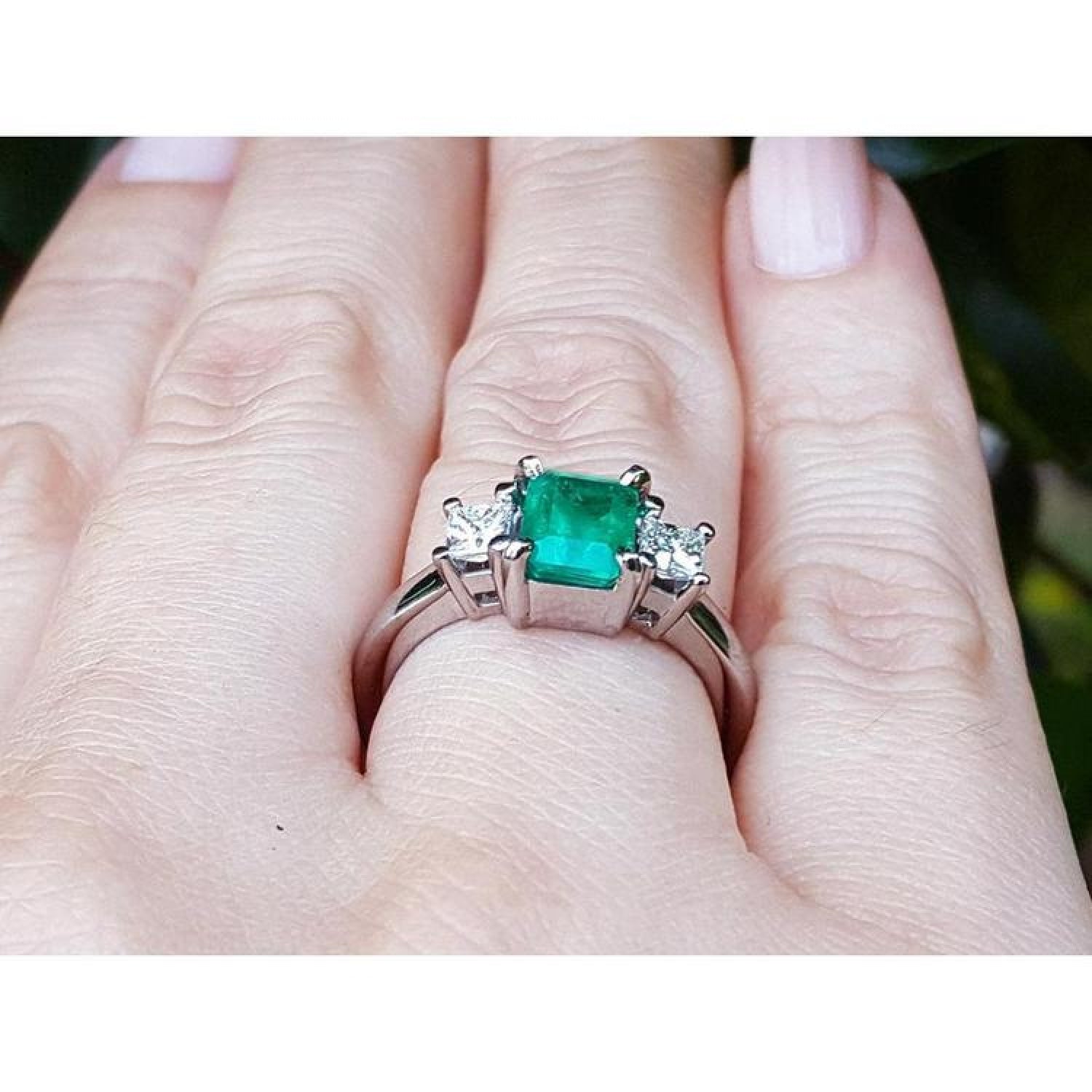 18k white gold three-stone ring with a vibrant emerald and two princess-cut diamonds on a hand.