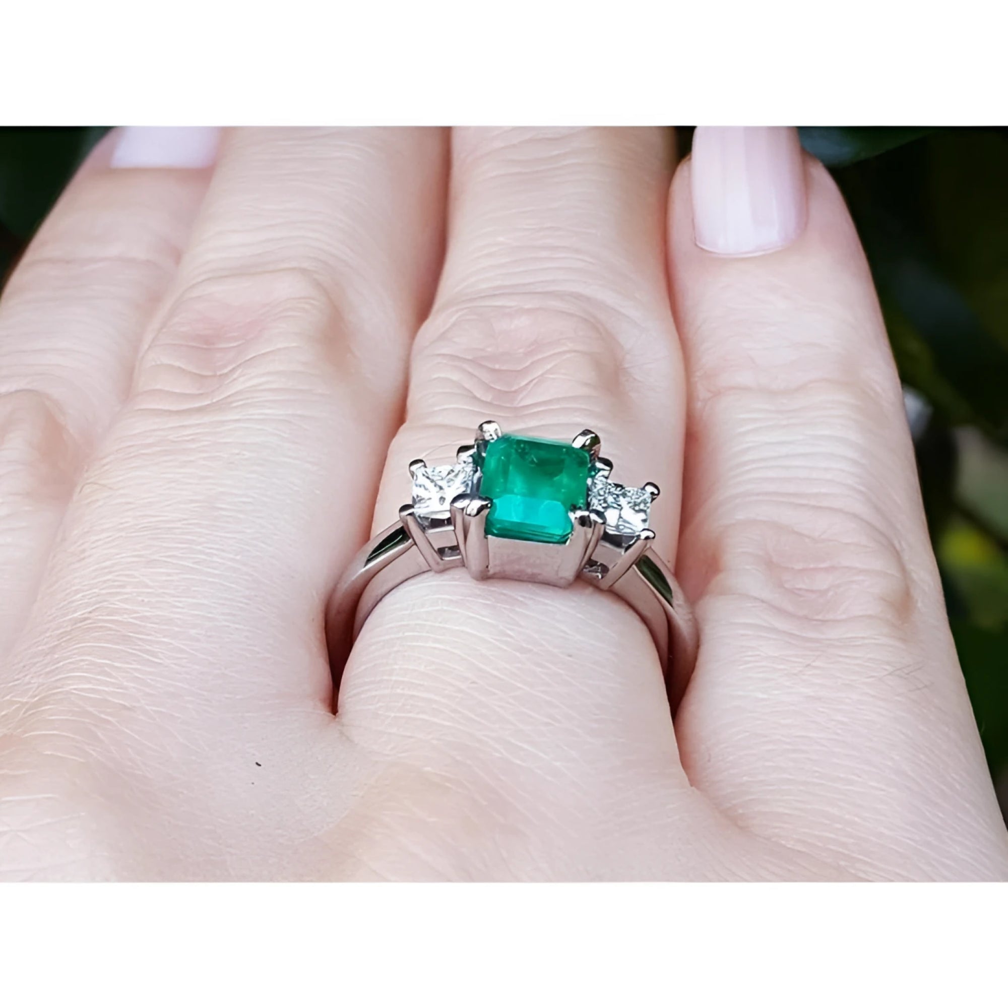 18k white gold three-stone ring with a vibrant emerald and two princess-cut diamonds on a hand.