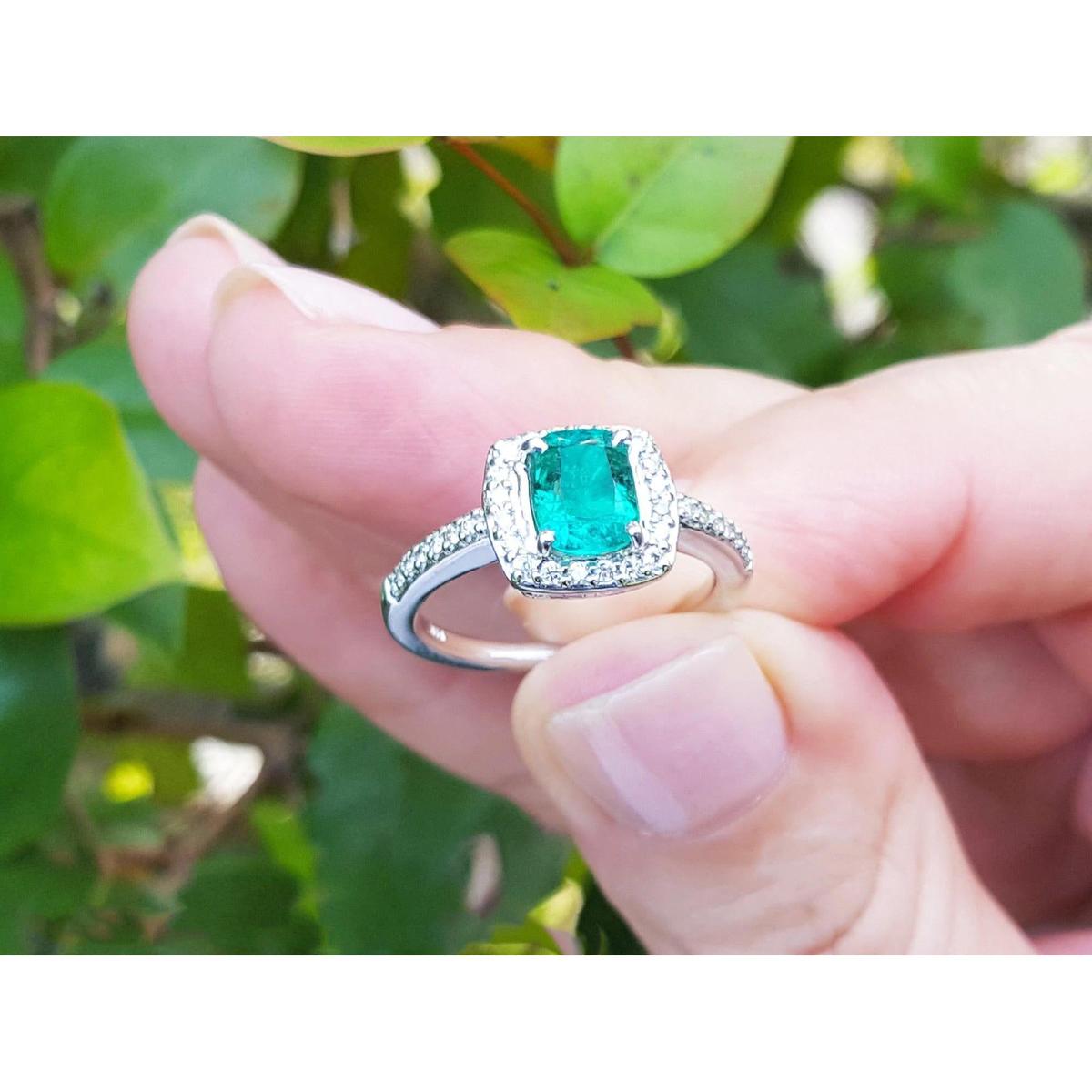 Emerald ring with diamond accents held between fingers against a green leafy background.