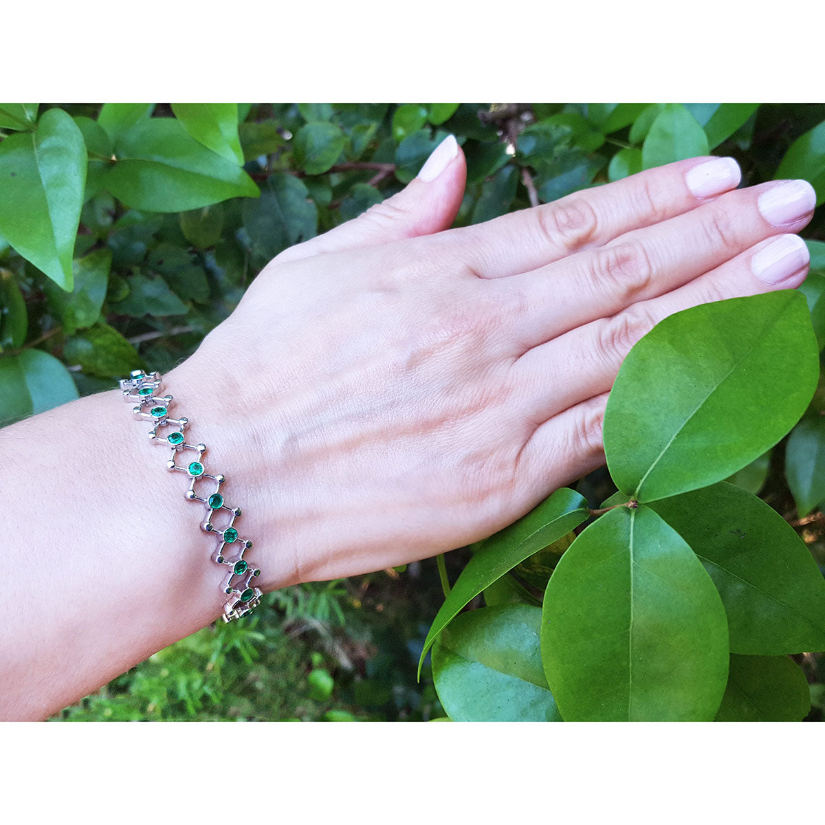 Hand wearing a white gold colombian emerald bracelet against a green leafy background.