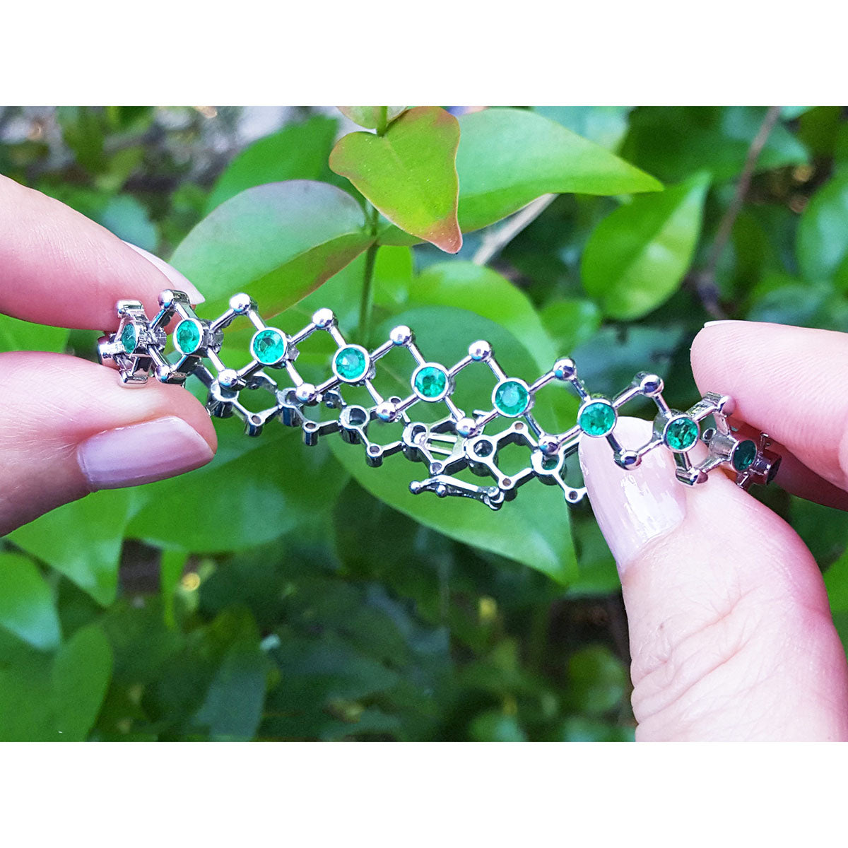 Whit gold bracelet with natural emeralds held between two fingers against a green leafy background.