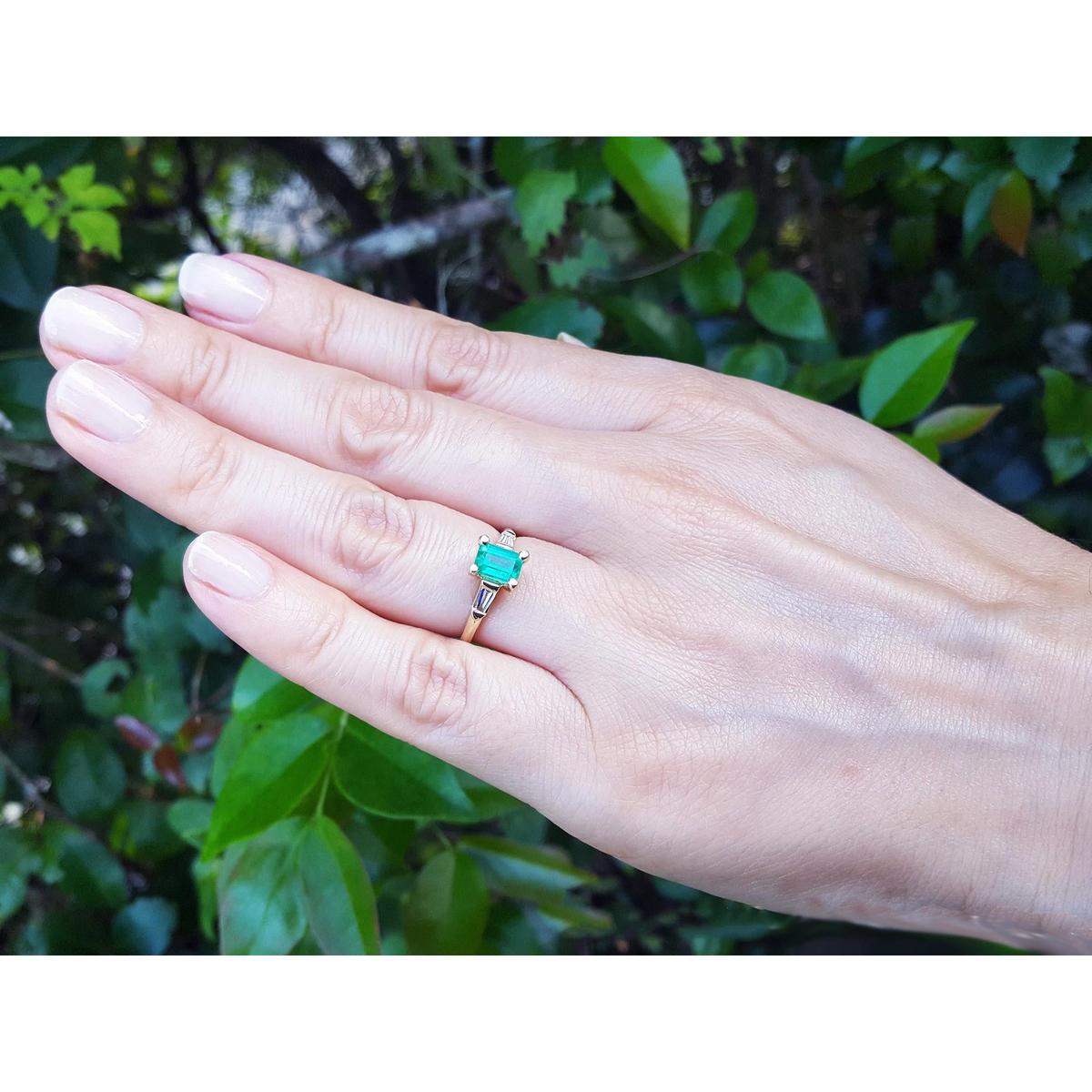 Hand wearing a natural emerald and baguette diamond ring against a natural background.