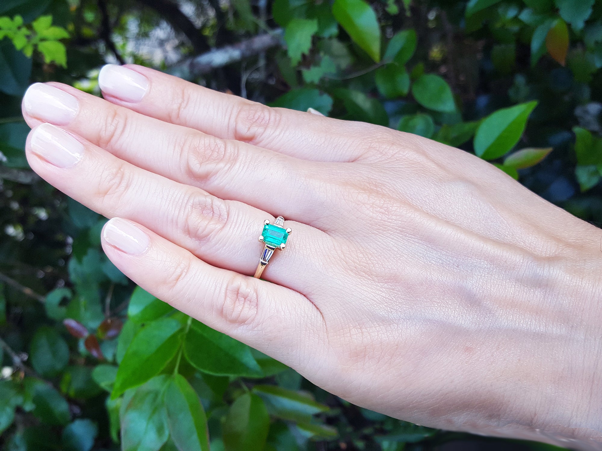 Hand wearing a natural emerald and baguette diamond ring against a natural background.