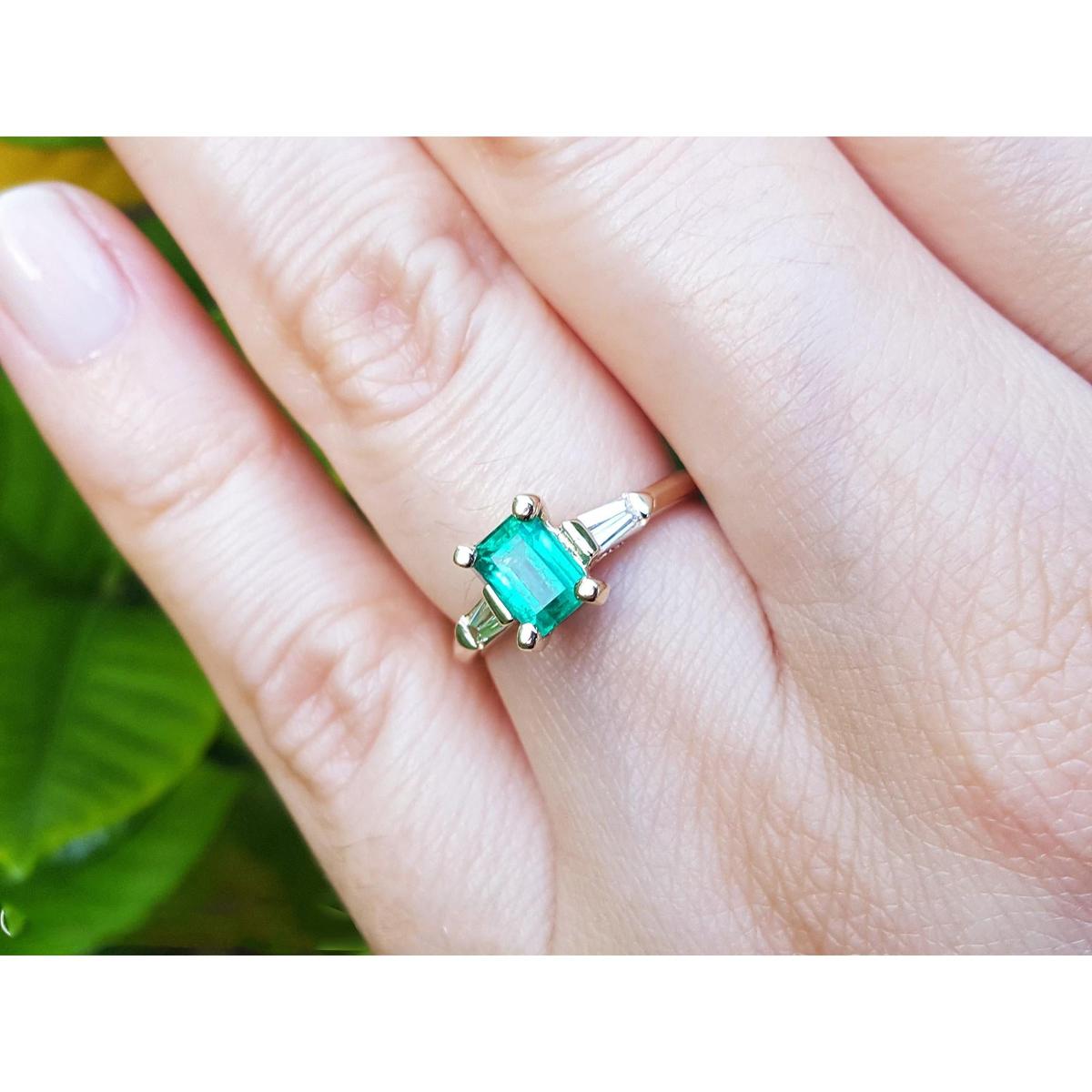 A gold ring with emerald and side baguette diamonds on a hand against a blurred green background.