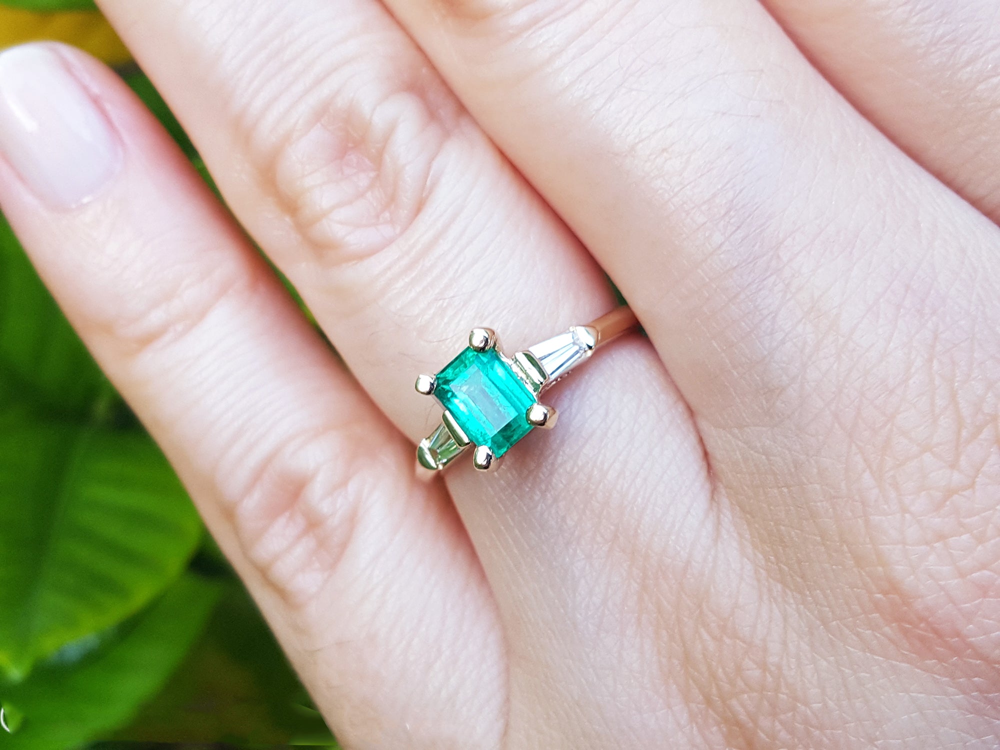 A gold ring with emerald and side baguette diamonds on a hand against a blurred green background.
