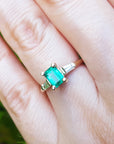 A gold ring with emerald and side baguette diamonds on a hand against a blurred green background.