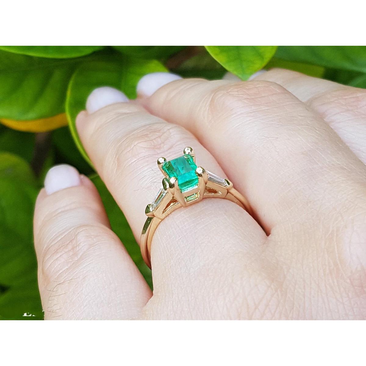 Yellow gold ring with emerald stone on a hand against a green leafy background.