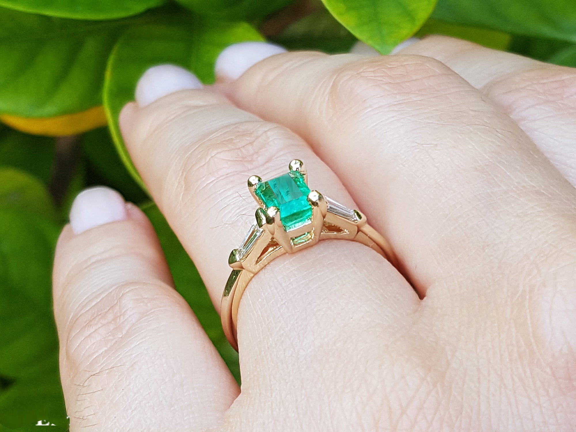 Yellow gold ring with emerald stone on a hand against a green leafy background.