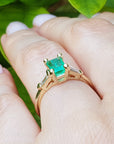 Yellow gold ring with emerald stone on a hand against a green leafy background.