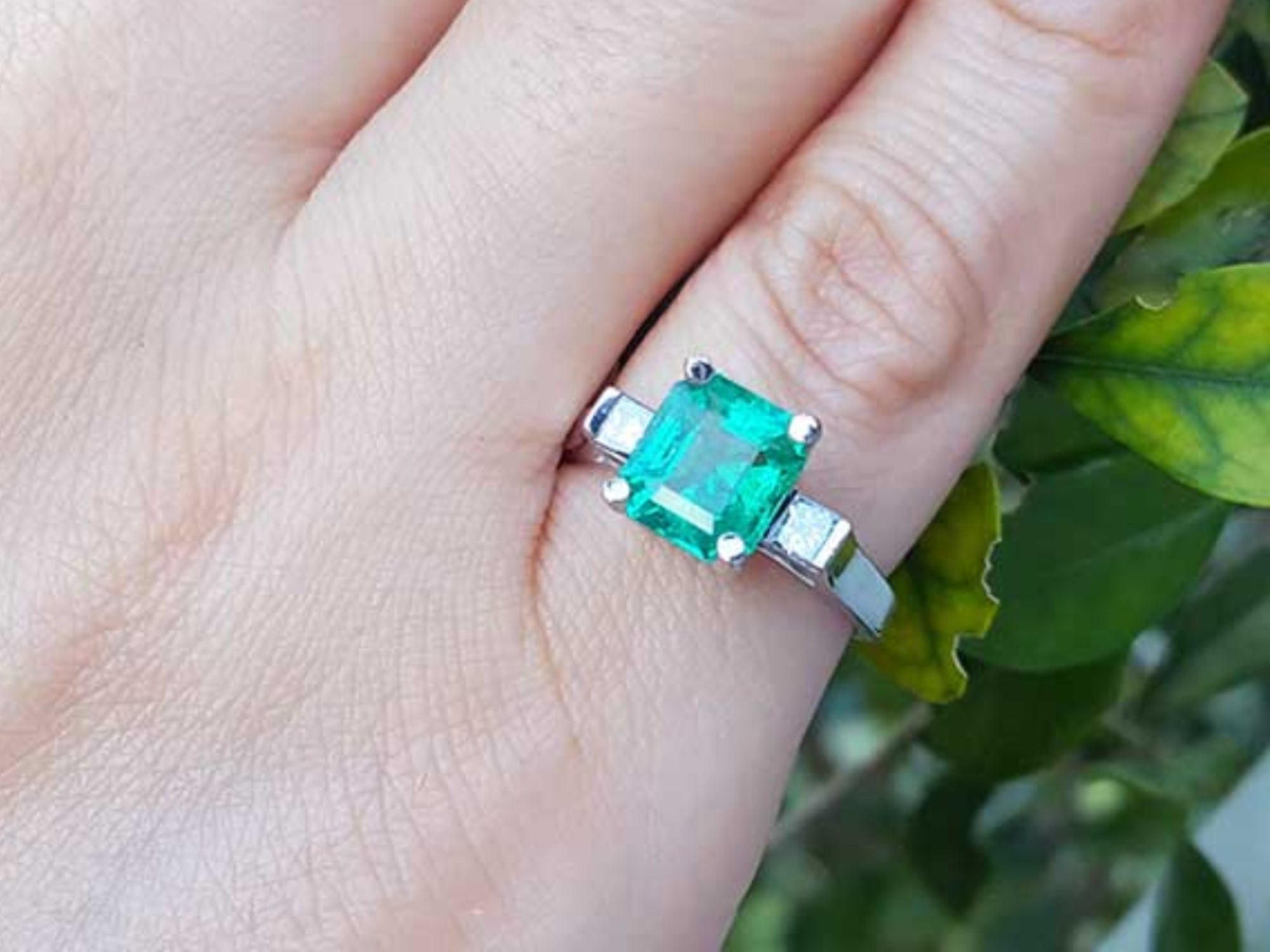 An emerald ring with side diamonds in white gold on a finger with a natural background.