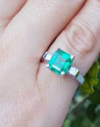 An emerald ring with side diamonds in white gold on a finger with a natural background.
