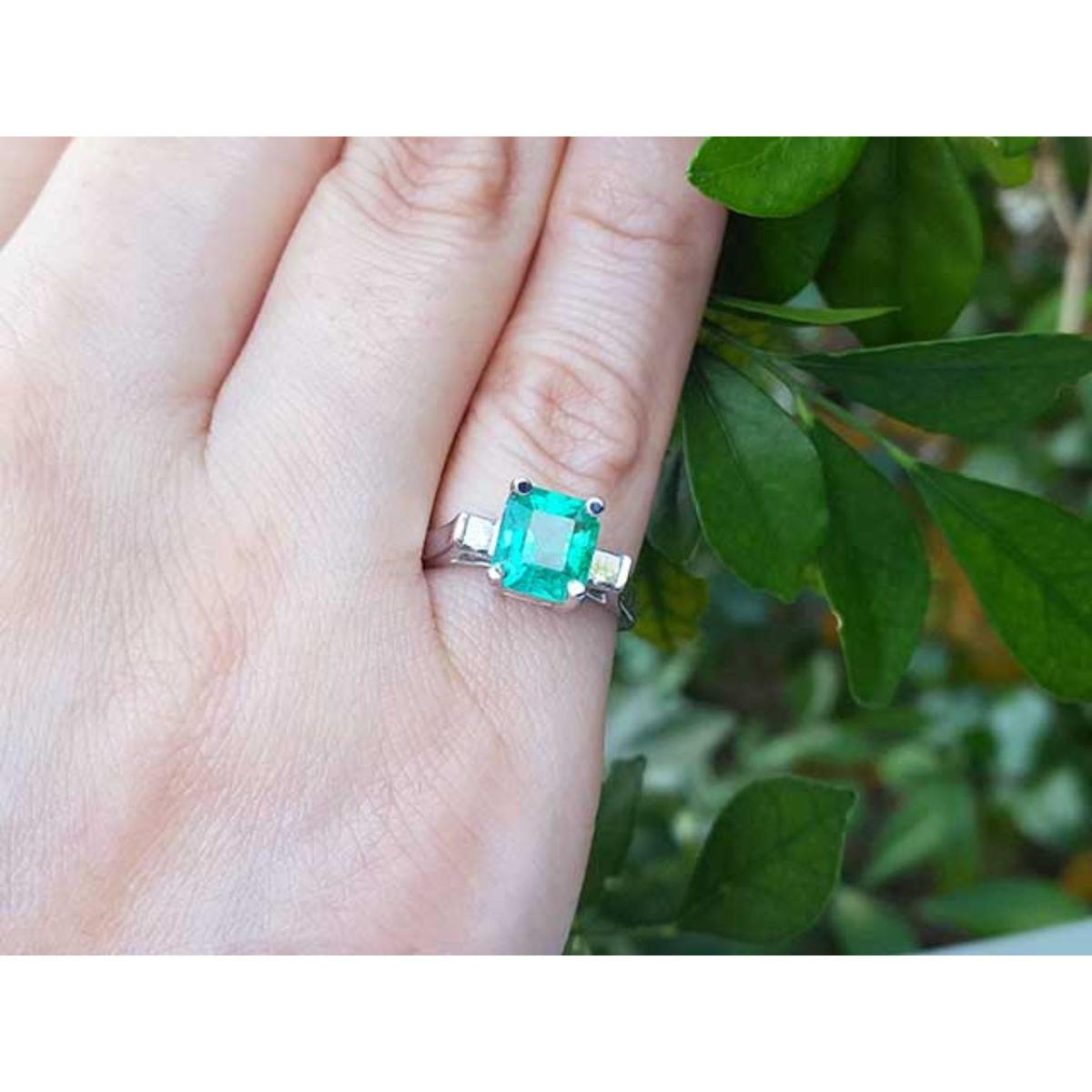 Emerald and side diamonds ring on a finger with a green leafy background.