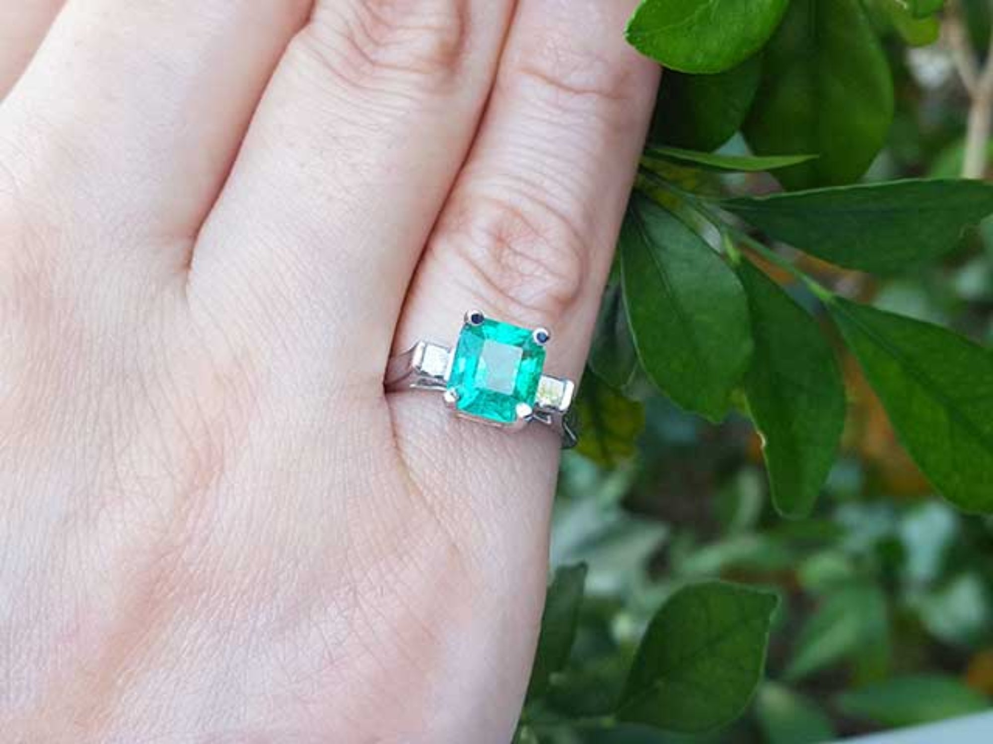 Emerald and side diamonds ring on a finger with a green leafy background.